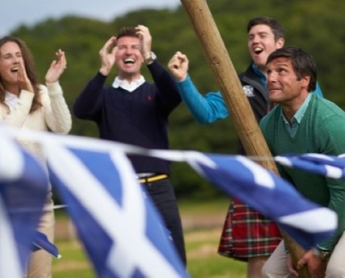 Man tossing a caber with three people cheering him on with Scottish flag bunting in the foreground